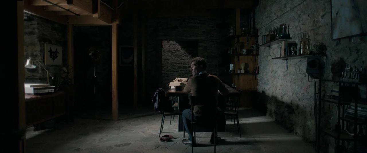 Oddity (2024) movie still: wide — A dimly lit room features rough stone walls and exposed wooden beams above. A man sits at a long table, turning…