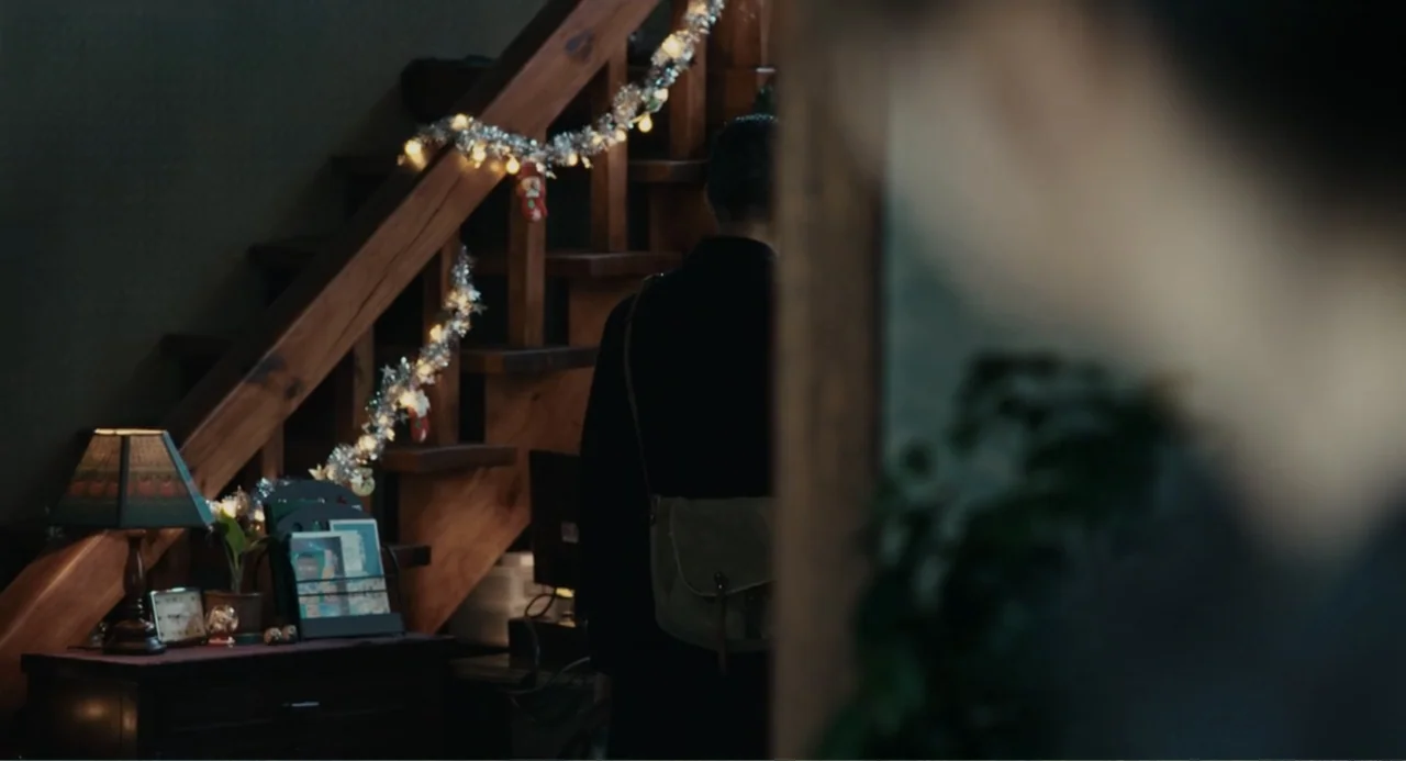 Winter in Sokcho (2025) movie still: medium — A dimly lit indoor space features a wooden staircase draped with silver tinsel lights. A person with a…