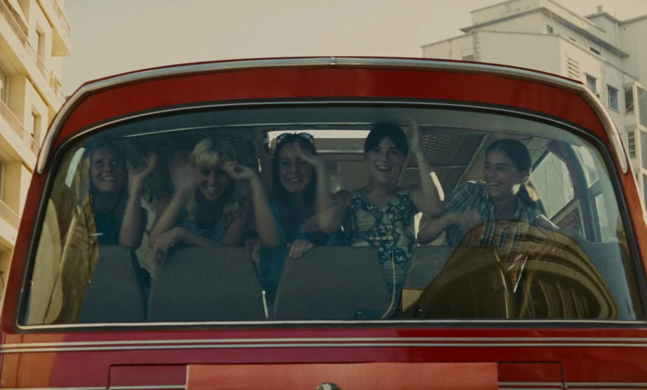 Z (1969) movie still: medium — A red bus travels along a street lined with light-colored multi-story apartment buildings. Five young women sit in…