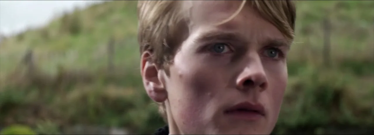 DannyBoy (2020) movie still: closeup — A grassy outdoor hillside serves as the backdrop for this close portrait. A young man stands in the open air…
