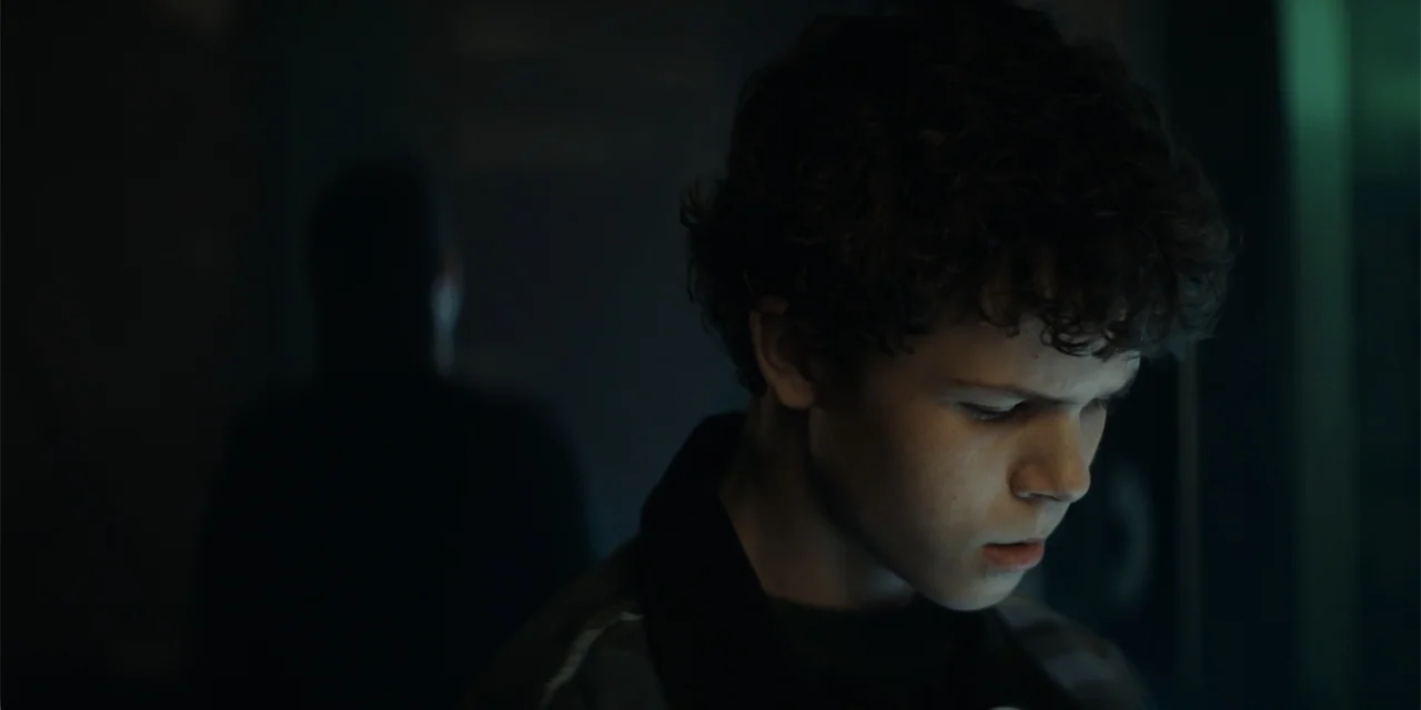 The Monkey (2025) movie still: closeup — A dark indoor space is illuminated by light emitting from a handheld electronic device. A youth stands in…