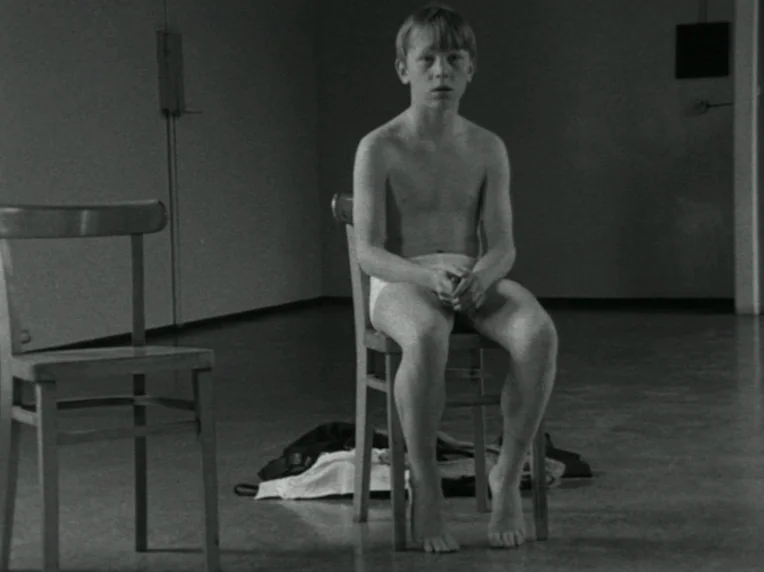 Children (1976) movie still: fullbody — A sparsely furnished room contains only two plain wooden chairs on a concrete floor. A young boy wearing…