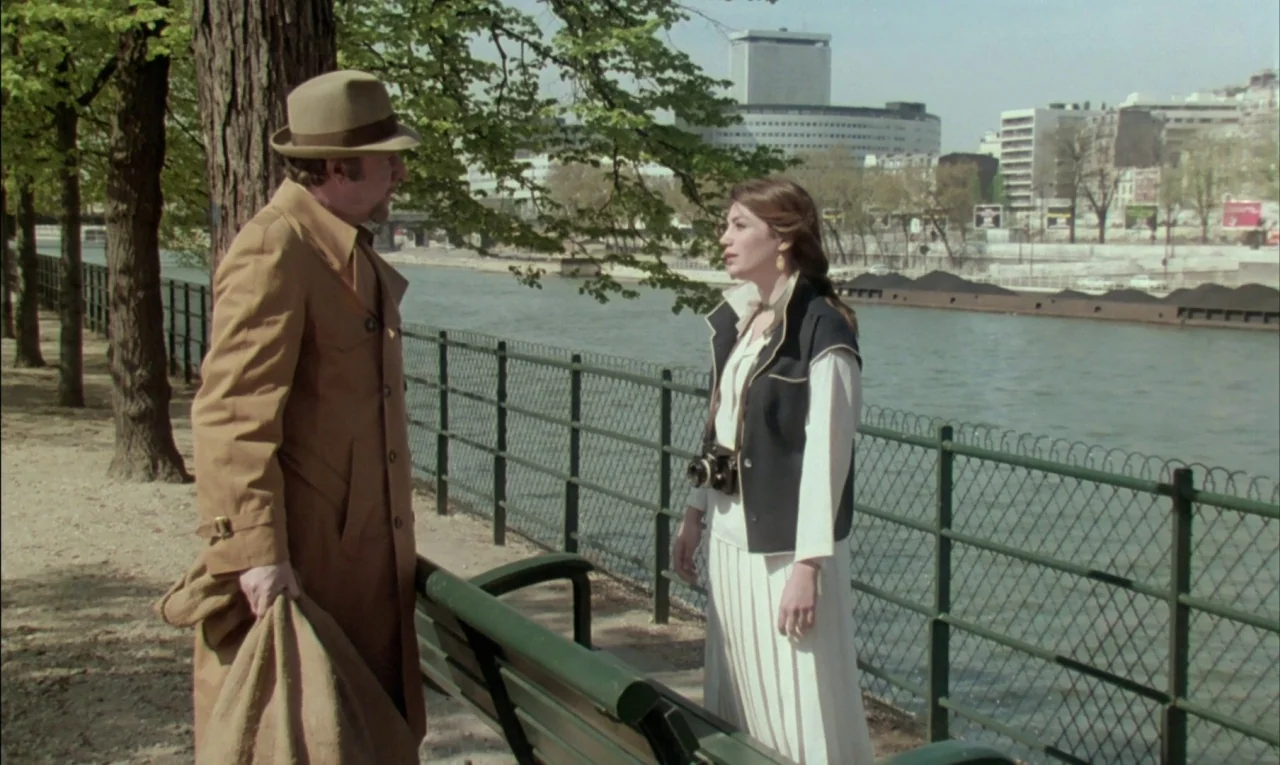 That Obscure Object of Desire (1977) movie still: fullbody — A riverside walkway in an urban setting with a metal fence and large trees. A man in a…