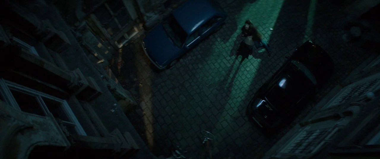 Atomic Blonde (2017) movie still: wide — A dark cobblestone courtyard framed by stone building walls at night. Two people stand in a concentrated…