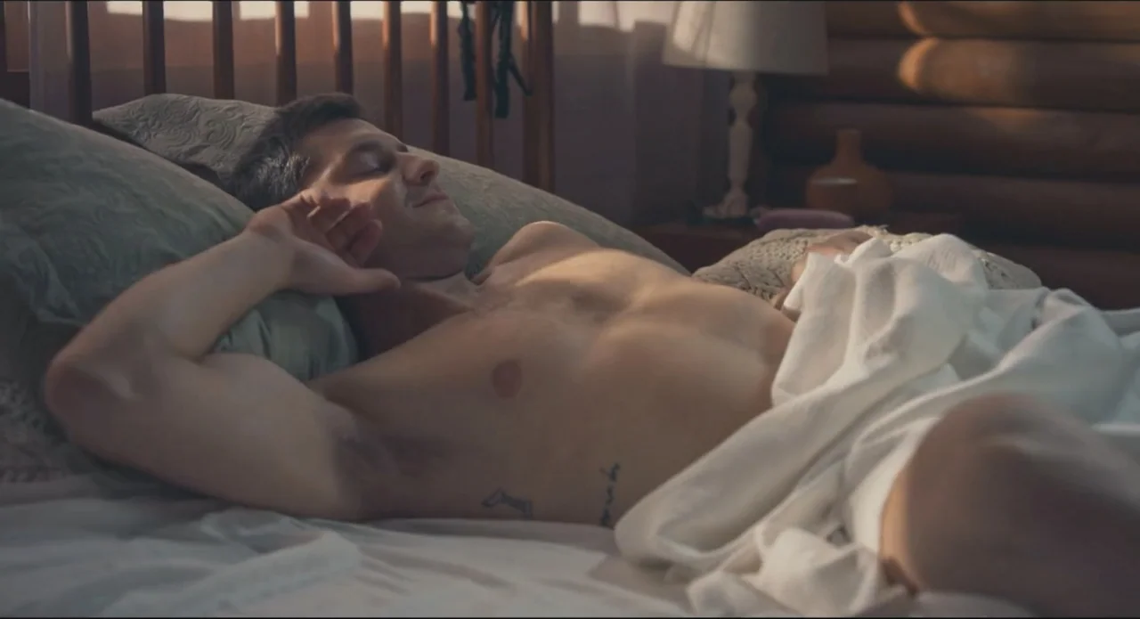 Proof of Love (2022) movie still: medium — A sunlit bedroom contains a bed with white linens and a wooden headboard. A shirtless young man lies…
