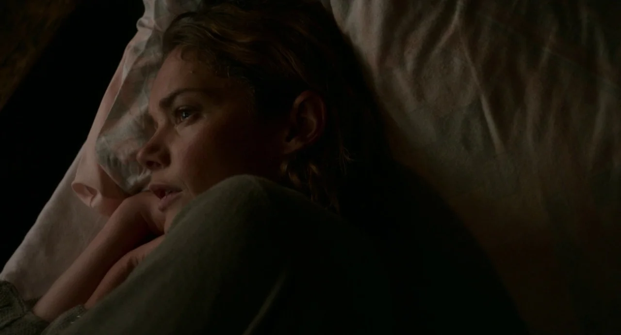 Dark River (2017) movie still: closeup — A person lies on a bed with patterned bedding in a dark room. The subject rests their head on a pillow…