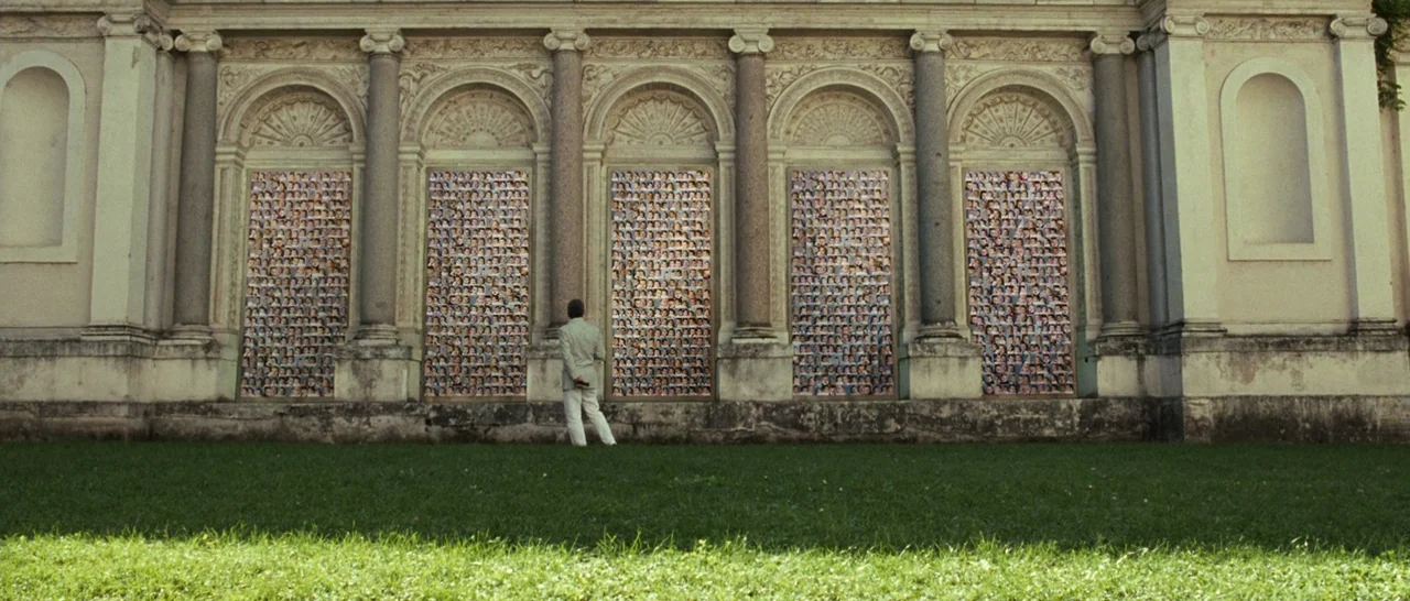 The Great Beauty (2013) movie still: wide — An ornate classical stone building facade features five arched niches filled with photo collages. A man…