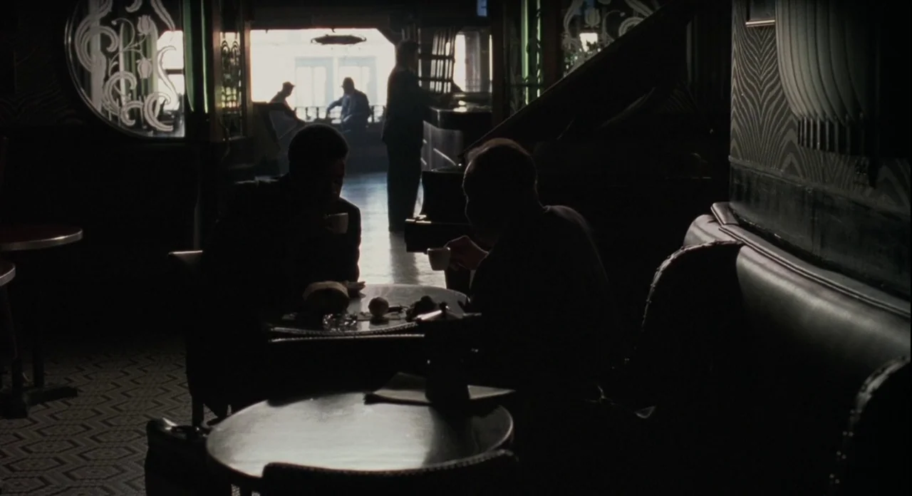 American Gangster (2007) movie still: wide — An interior cafe space features art deco style wall patterns and large windows. Two men sit in…