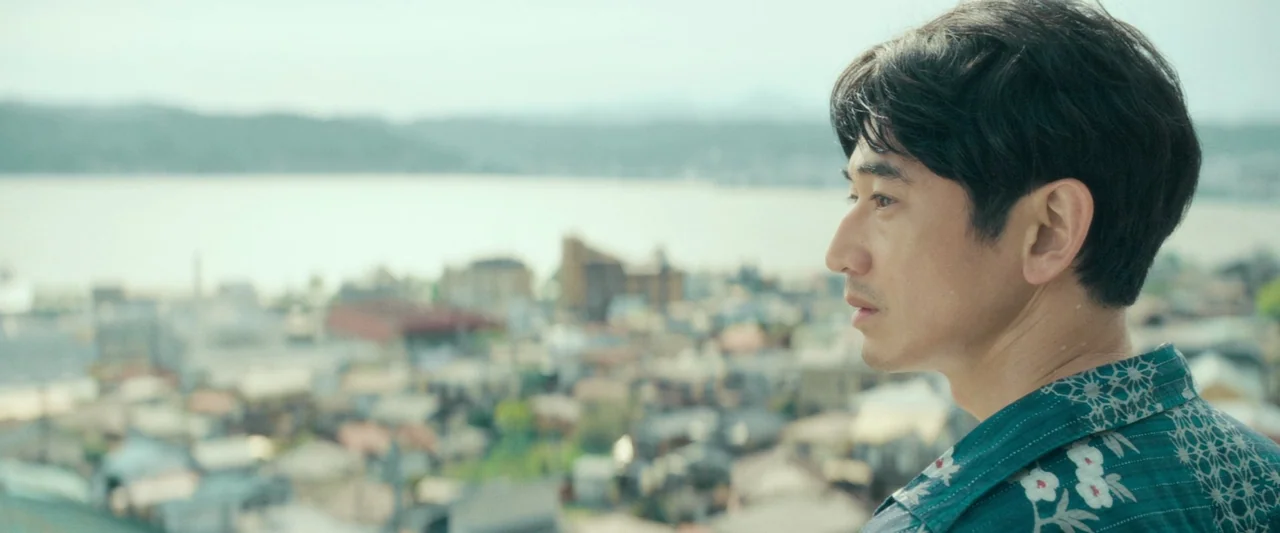 Monster (2023) movie still: medium — A man stands on an overlook viewing a dense seaside town in the distance. He gazes off-frame toward the water…