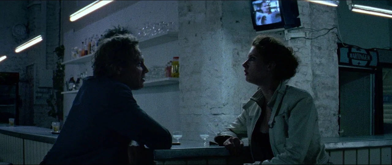 Luz (2018) movie still: medium — A dimly lit room with a metallic counter and white textured brick walls. A man and a woman sit facing each other…