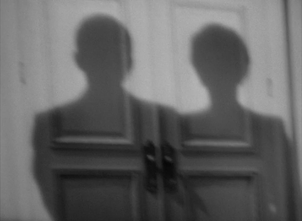 Notorious (1946) movie still: medium — A pair of dark shadows are cast onto a multi-paneled wooden interior door. The silhouettes of a man and woman…