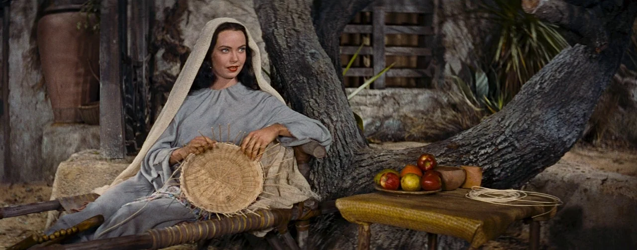 The Robe (1953) movie still: medium — A woman sits beneath a large tree in a rustic, arid environment. She holds a circular woven basket in her lap…