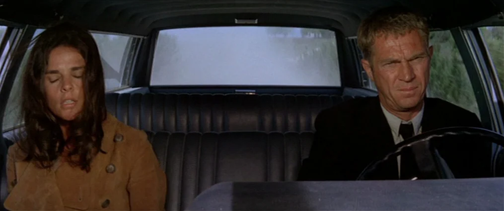 The Getaway (1972) movie still: medium — A man and a woman sit in the front seat of a moving vehicle. The car interior features black ribbed vinyl…
