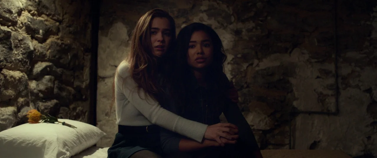 Split (2016) movie still: medium — A dark stone-walled cellar room features a single bed with a white pillow. Two young women sit side by side on…