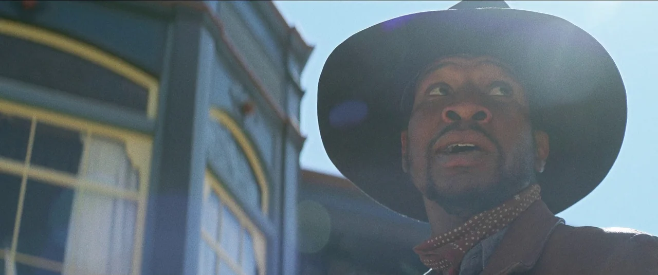 The Harder They Fall (2021) movie still: closeup — A wooden exterior of a building sits in the sun behind a man. The man in a wide-brimmed hat gazes…