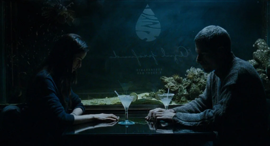 Sidewalls (2011) movie still: medium — A man and a woman sit at a table in front of a large, illuminated aquarium. The restaurant is dimly lit,…
