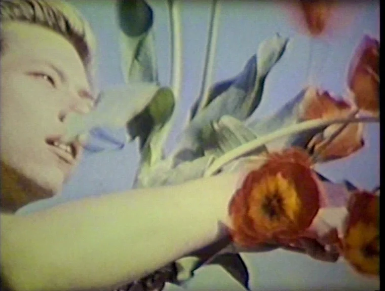 The Angelic Conversation (1985) movie still: closeup — A man holds a bouquet of orange tulips against a clear bright sky. The low-resolution footage…