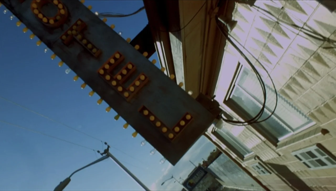 Dark Country (2009) movie still: establishing — An exterior view of a motel building with a prominent illuminated lightbulb sign. The camera is…