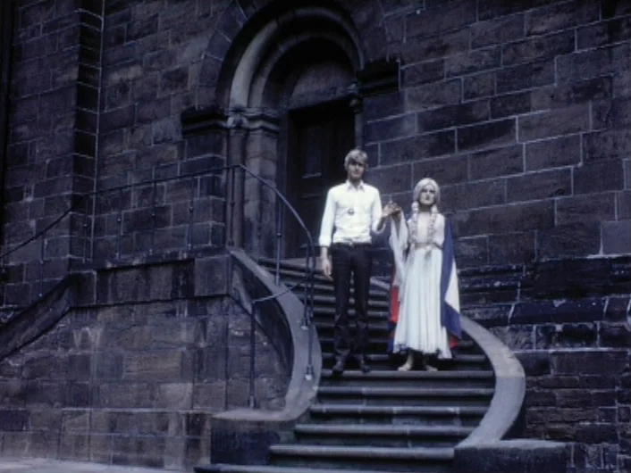 Eika Katappa (1969) movie still: fullbody — An imposing dark stone building features a curved exterior staircase with ornate iron railings. A young…