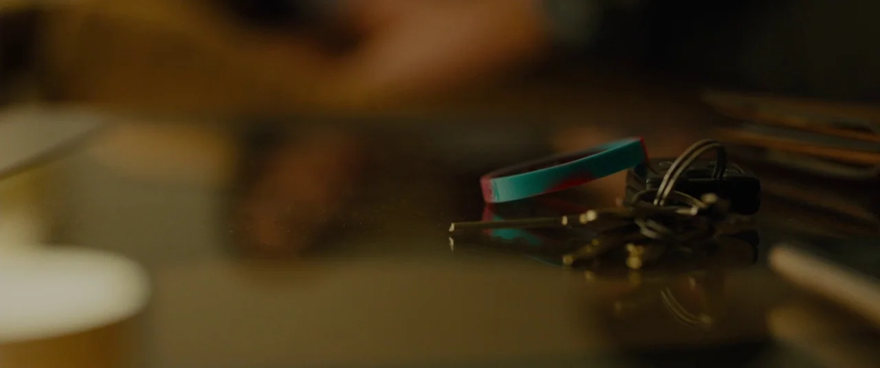 Sicario (2015) movie still: closeup — A set of keys and a silicone wristband rest on a dark reflective surface. The background is blurred and out of…