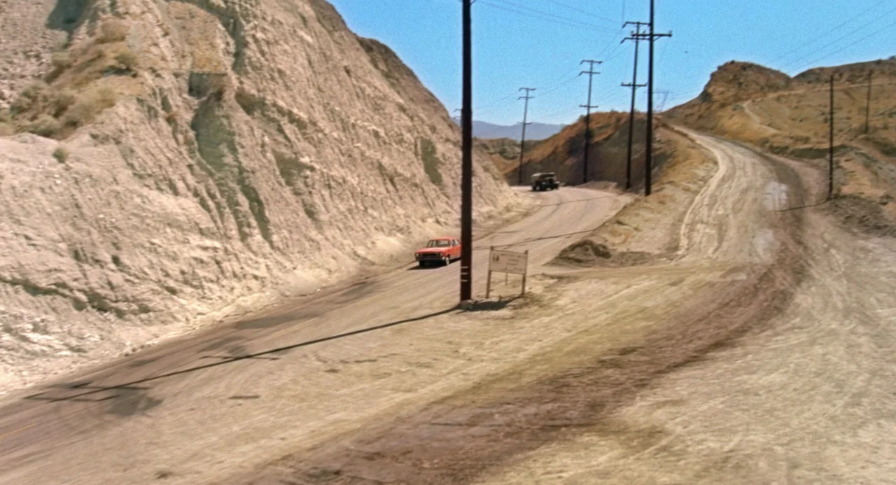 Duel (1971) movie still: wide — An arid, hilly landscape features a dusty dirt road winding through rocky terrain. A single red sedan drives along…
