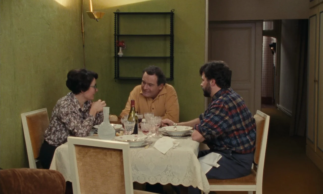 A Tale of Winter (1992) movie still: medium — A dining area features olive green walls, a lace tablecloth, and simple wooden chairs. Three people…