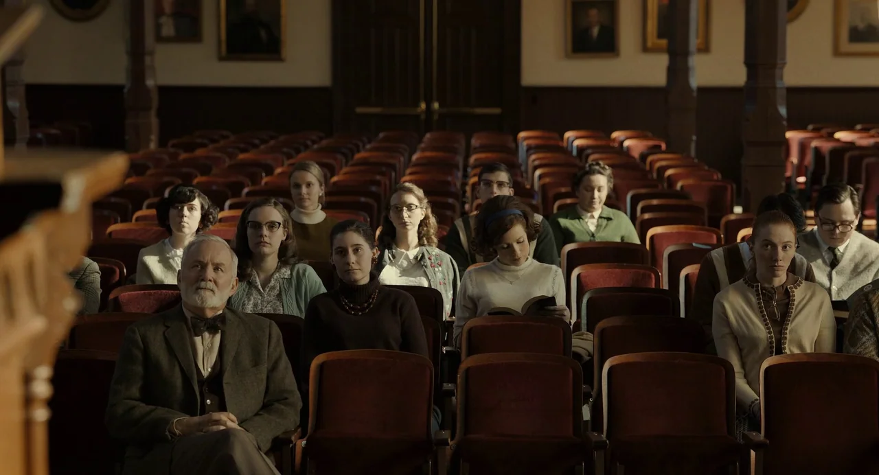 The Eyes of Tammy Faye (2021) movie still: wide — A dimly lit auditorium features rows of vintage red velvet chairs filled with people. The audience…