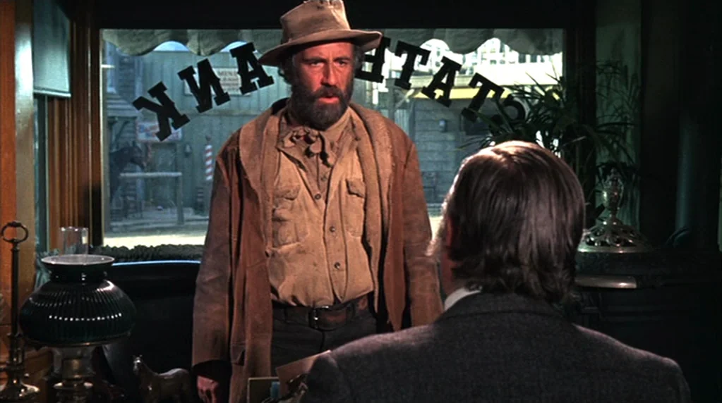 The Ballad of Cable Hogue (1970) movie still: medium — A dimly lit room with a large front window looking out onto a street. A bearded man stands…