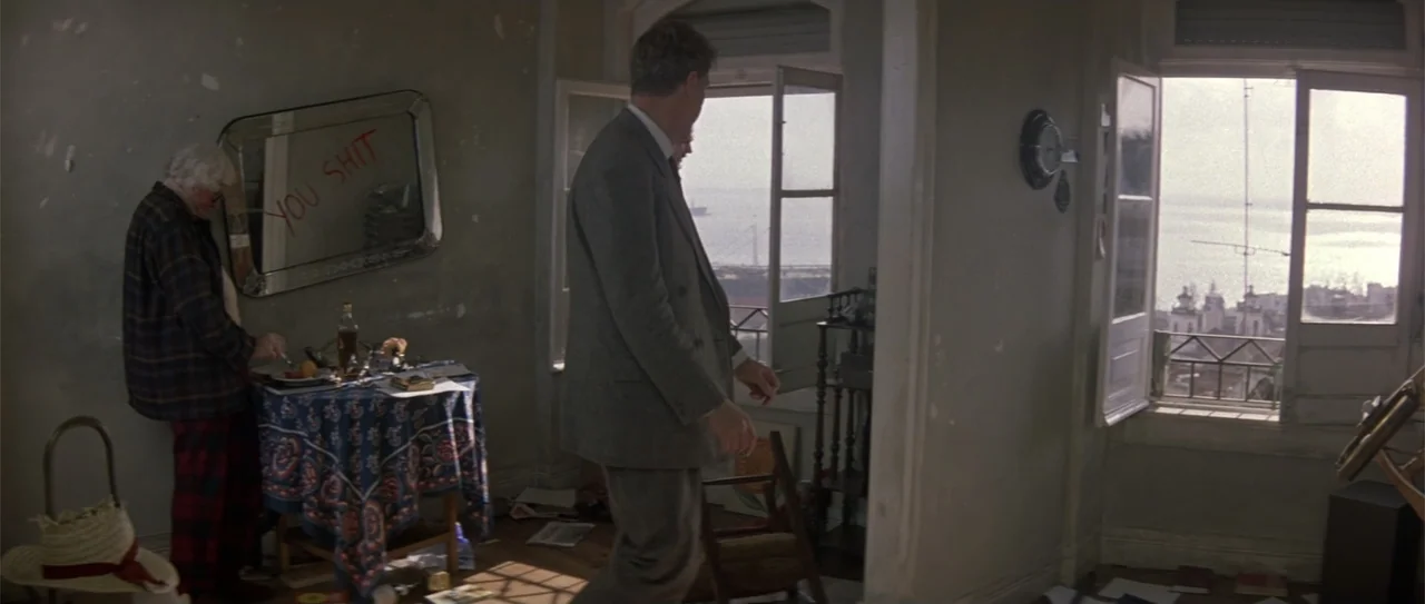 The Russia House (1990) movie still: fullbody — An apartment room contains a table with a blue patterned cloth and wall mirror. A man in a suit…