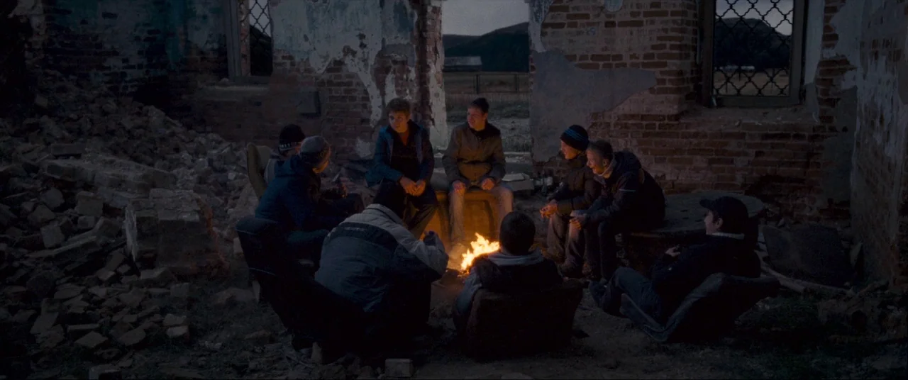 Leviathan (2014) movie still: wide — A group of teenagers sits in a circle inside a ruined brick building shell. They are gathered around a small,…