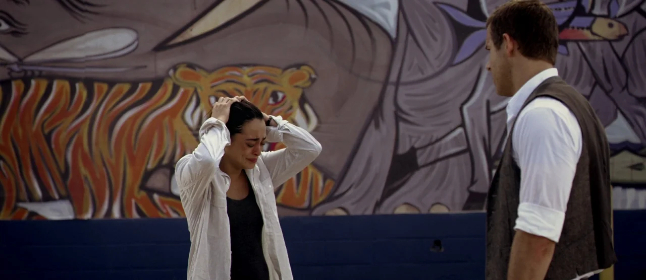 Self/less (2015) movie still: medium — A mural depicting large tigers serves as the backdrop to this outdoor area. A woman clutching her head stands…