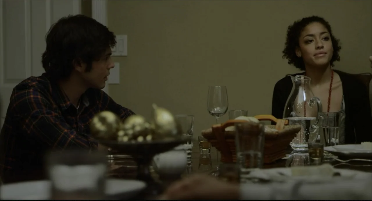 Freedom (2024) movie still: medium — A residential dining room at night contains a dark wooden table with place settings. A young man and woman sit…