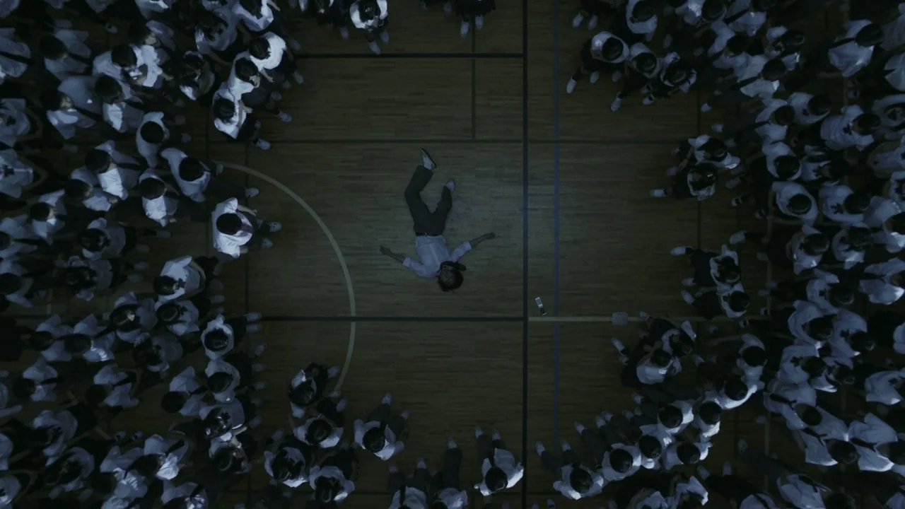 Confessions (2010) movie still: wide — A high-angle view looking down at a large school gymnasium with wood floors. A lone student lies flat in the…