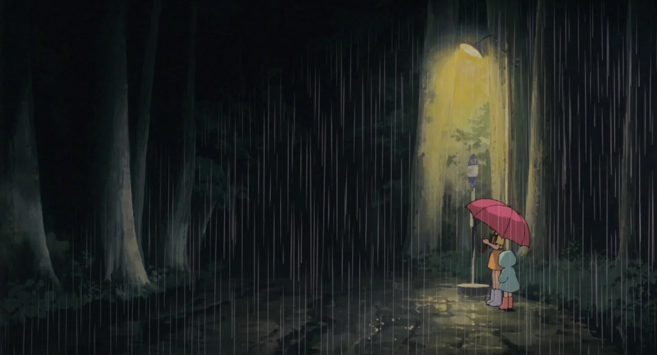 My Neighbour Totoro (1988) movie still: wide — A dark, wet wooded area with towering trees stands in a heavy downpour at night. Two people stand…