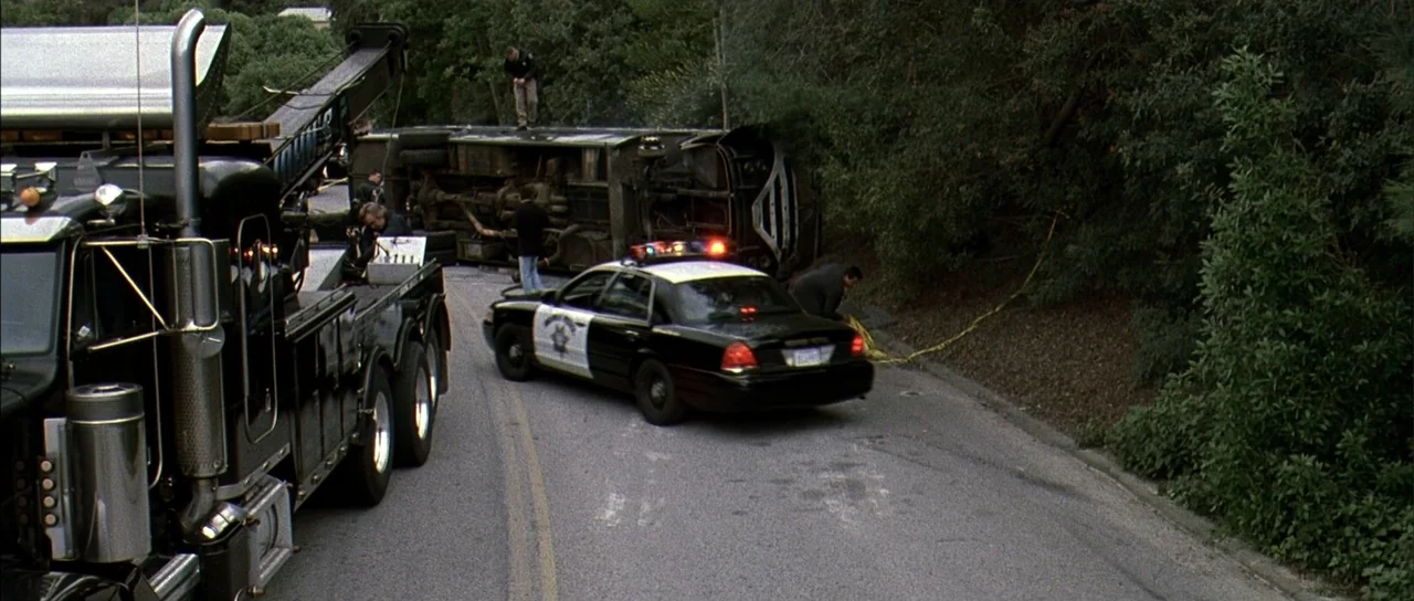 A Man Apart (2003) movie still: wide — A winding asphalt road passes through a wooded area with dense greenery. A large bus lies overturned on its…