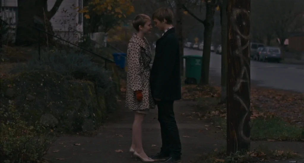 Restless (2011) movie still: fullbody — A quiet suburban residential sidewalk during late autumn in the fading evening light. A young couple stands…