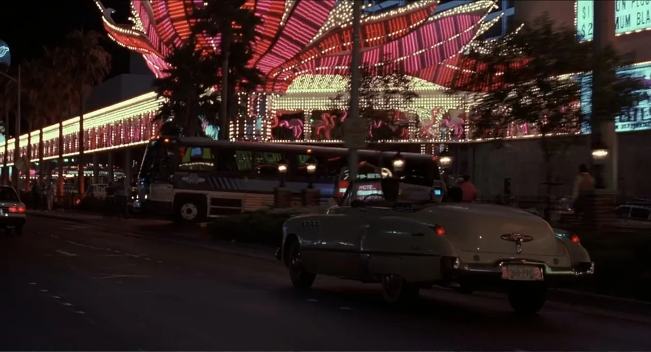 Rain Man (1988) movie still: wide — A Las Vegas street at night features bright pink flamingo neon lights on a facade. A classic white convertible…