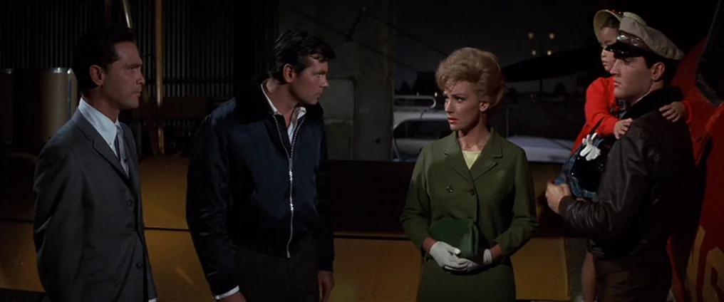 It Happened At The World’s Fair (1963) movie still: medium — An open-air aircraft hangar at night shelters four adults and a small child. The group…