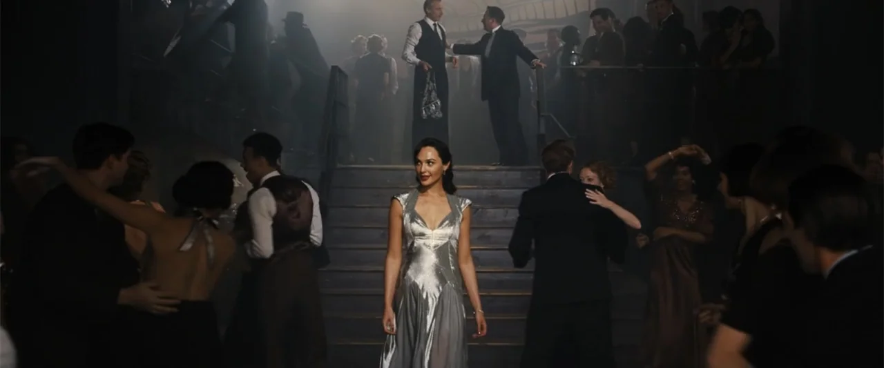 Death on the Nile (2022) movie still: fullbody — A dark, smoky ballroom with wooden stairs and numerous figures dressed in formal 1940s evening…