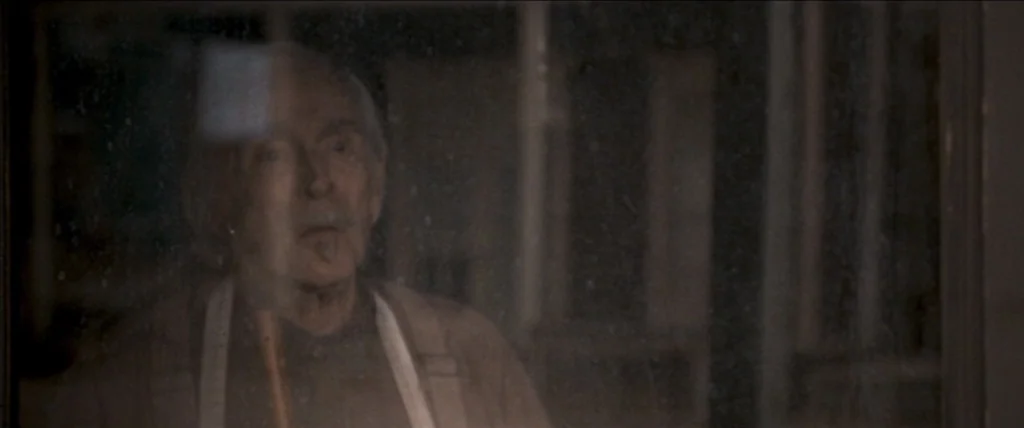In A Valley of Violence (2016) movie still: medium — An elderly man peers through a dusty, dark glass window from a dimly lit interior. The glass is…