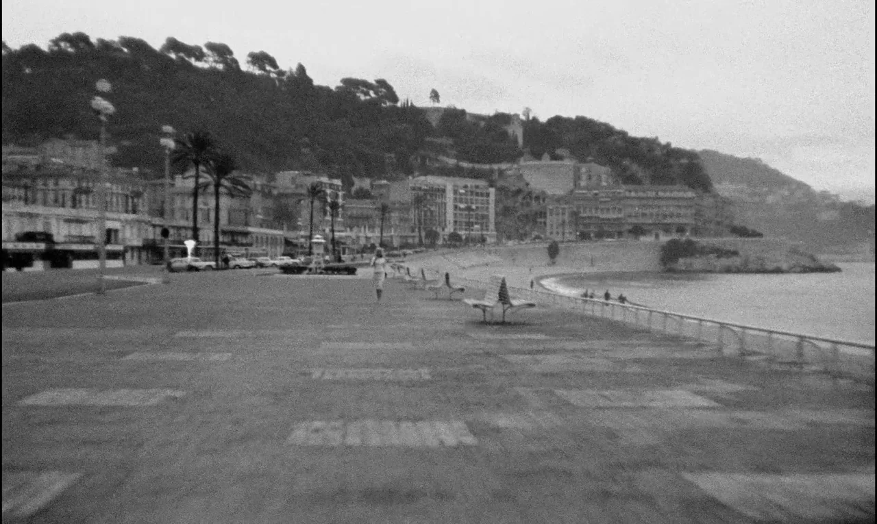 Bay of Angels (1963) movie still: wide — A wide view of the Promenade des Anglais with palm trees and urban buildings behind. A lone woman walks…