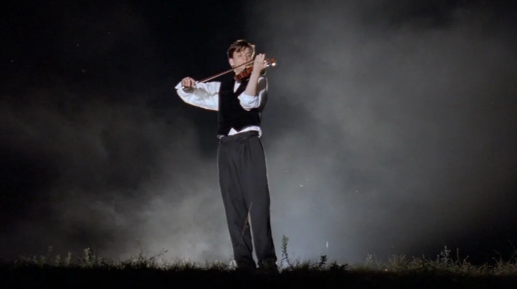 Malina (1991) movie still: fullbody — A man stands alone in a dark, hazy field under an obscured night sky. He plays a violin while positioned…