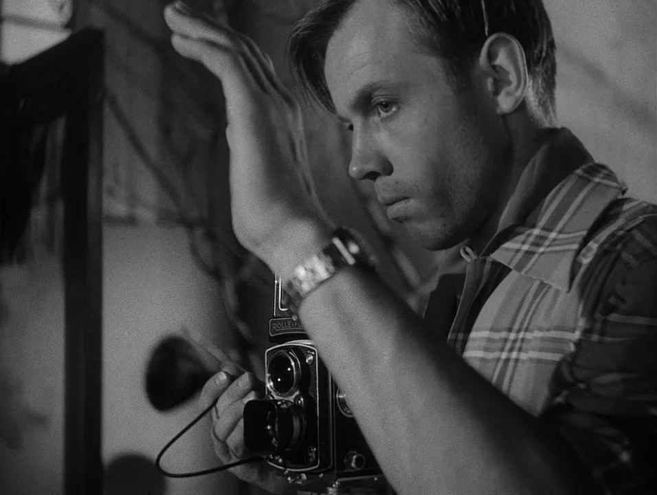 Dreams (1955) movie still: medium — A dimly lit room serves as a workspace for a person using equipment. A young man wearing a plaid shirt holds a…