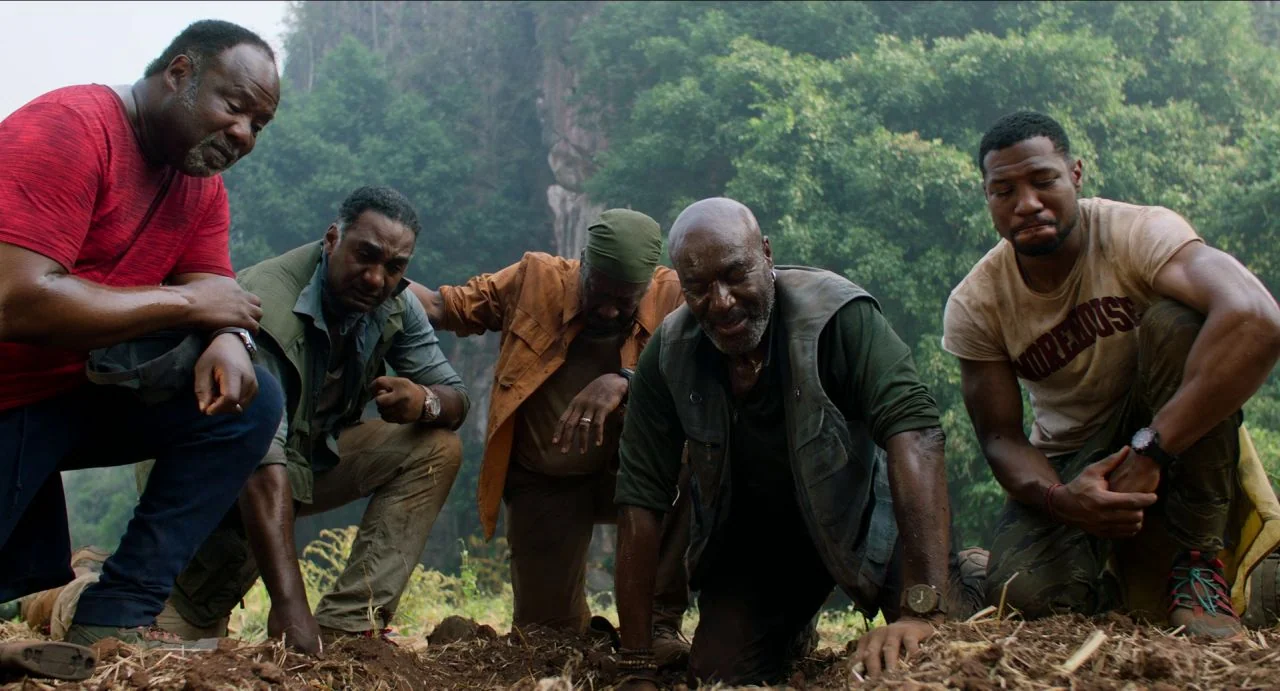 Da 5 Bloods (2020) movie still: medium — A group of five men crouch together in a muddy jungle clearing. They examine the earth with expressions of…