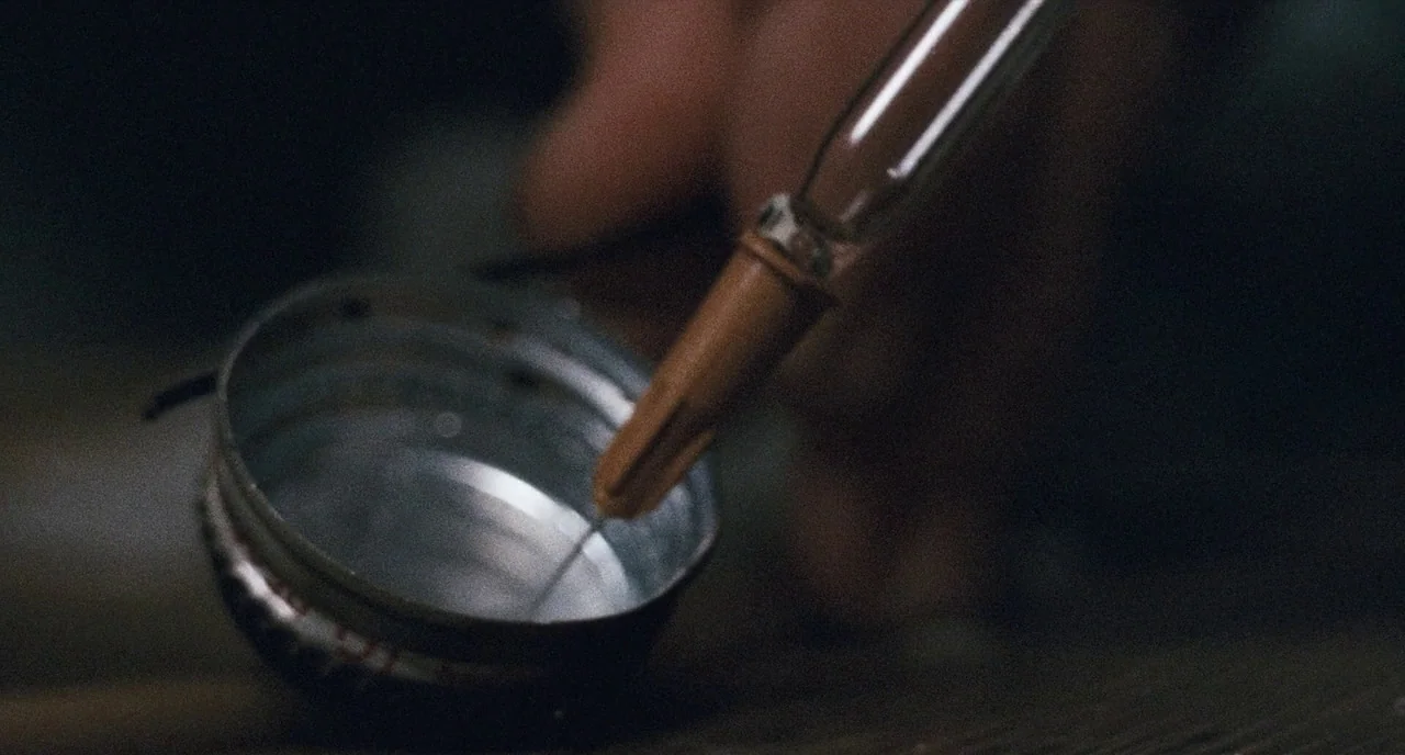 The Panic in Needle Park (1971) movie still: closeup — A close-up view of a metal bottle cap filled with clear liquid. A syringe needle dips into…