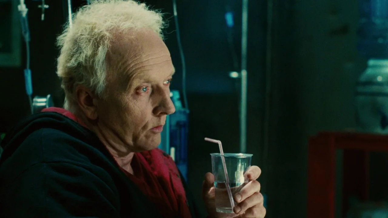 Saw II movie still: closeup — A dimly lit room with medical equipment stands in the background shadows. The man holds a clear plastic cup while…
