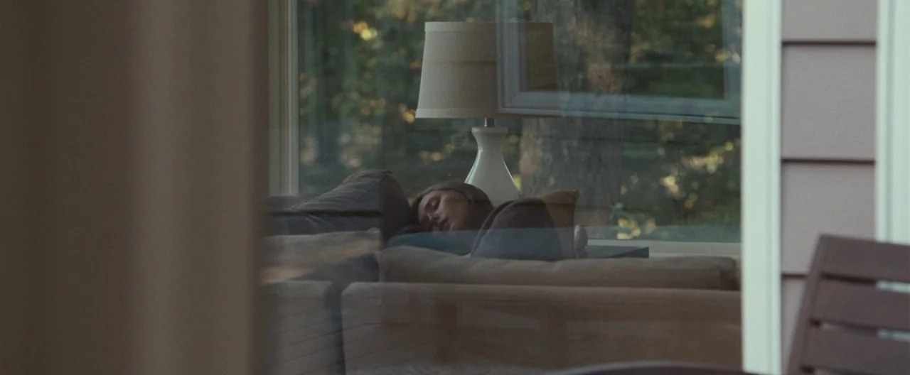 Martha Marcy May Marlene (2011) movie still: wide — A view through a window reveals a quiet interior with a person sleeping on furniture. A lamp…