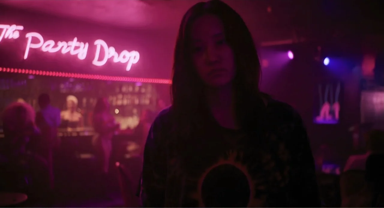 Mona Lisa and the Blood Moon (2022) movie still: medium — A dark nightclub interior features a bright pink glowing 'The Party Drop' neon sign. A…