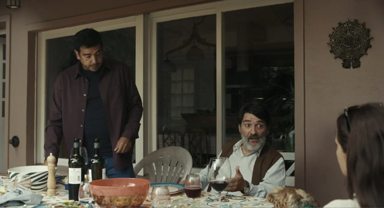 Belén (2025) movie still: medium — A residential outdoor patio features a dining table covered in dishes and wine bottles. Two men converse while…