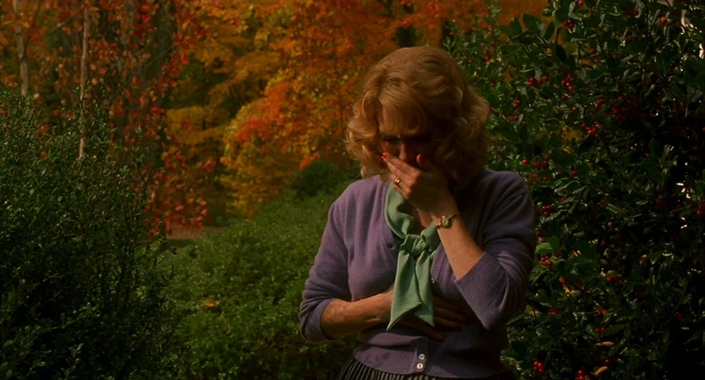Far From Heaven (2002) movie still: medium — A woman stands in a lush garden filled with autumn leaves and green foliage. She holds her hand to her…